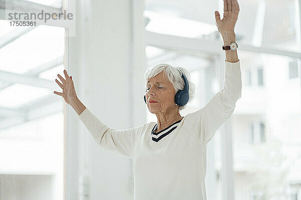 Ältere Frau mit erhobenen Händen  die zu Hause Musik über Kopfhörer hört