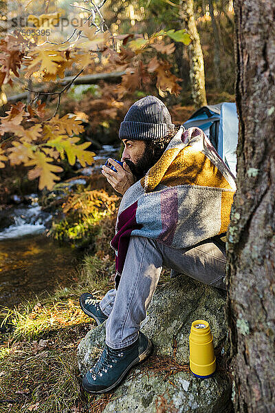 Mann trinkt Kaffee und sitzt auf einem Felsen im Herbstwald
