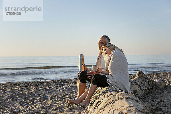 Paar sitzt auf einem Baumstamm und genießt den Sonnenuntergang am Strand