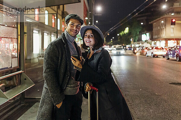 Lächelndes junges Paar mit Smartphone und Rollgepäck in der Stadt