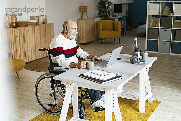 Leitender Geschäftsmann im Rollstuhl mit Kaffeetasse und Laptop im Heimbüro