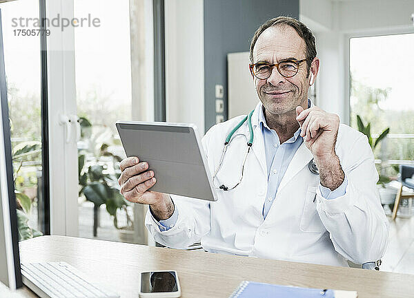 Lächelnder Arzt mit Tablet-PC am Schreibtisch im Krankenhaus