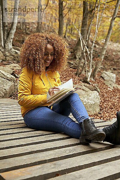 Junge Frau mit lockigem Haar liest Buch auf Brücke im Herbstwald