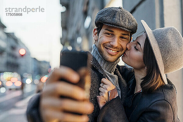 Junge Frau küsst ihren glücklichen Freund  während sie ein Selfie mit dem Smartphone macht