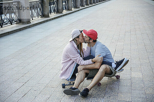 Liebevolles Paar  das sich küsst und auf dem Skateboard am Fußweg sitzt