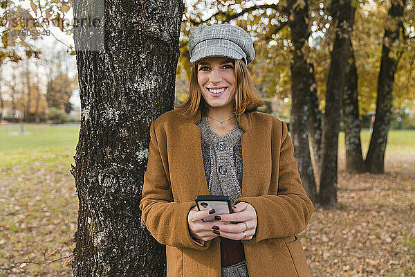 Lächelnde Frau mit Smartphone lehnt an Baumstamm