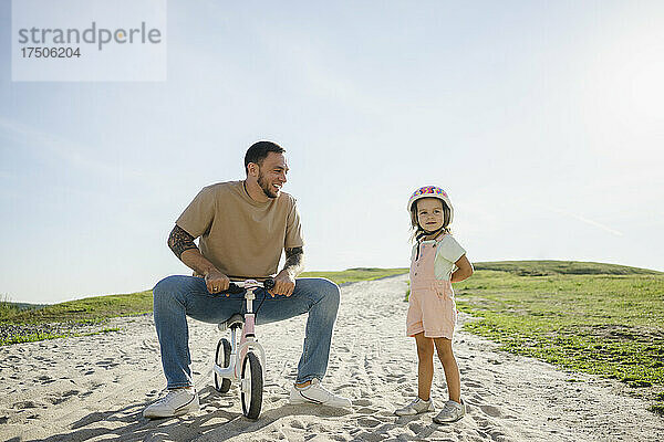 Verspielter Vater sitzt auf dem Fahrrad und schaut seine Tochter an  die im Sand steht