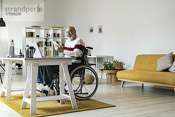 Älterer behinderter Geschäftsmann mit Mobiltelefon und Laptop im Heimbüro