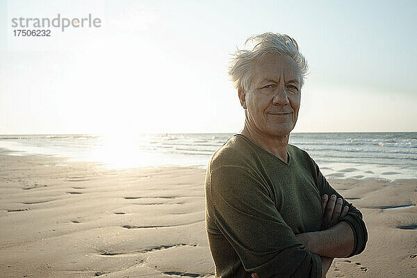 Älterer Mann lächelt mit verschränkten Armen am Strand