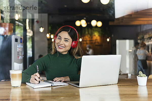 Lächelnde Geschäftsfrau mit kabellosen Kopfhörern und Laptop im Café