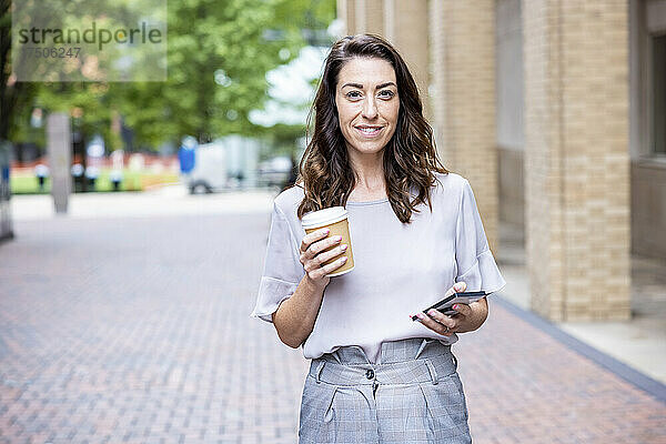 Lächelnde Geschäftsfrau hält Einwegbecher und Smartphone auf dem Fußweg