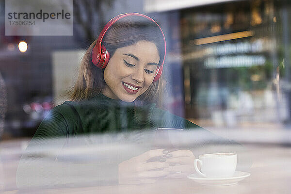 Lächelnde Frau mit kabellosen Kopfhörern  die im Café ihr Mobiltelefon benutzt