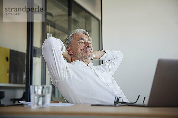 Lächelnder Geschäftsmann entspannt sich mit den Händen hinter dem Kopf im Büro