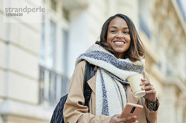 Lächelnde Frau mit Schal  Einwegbecher und Mobiltelefon in der Hand