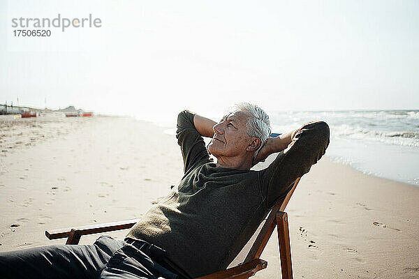 Älterer Mann mit den Händen hinter dem Kopf entspannt sich auf einem Stuhl am Strand