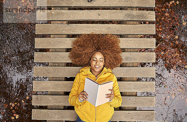 Sorglose Frau liegt auf einer Brücke und liest ein Buch im Herbstwald
