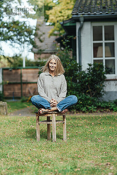 Lächelnde Frau sitzt mit gekreuzten Beinen auf einem Hocker im Hinterhof