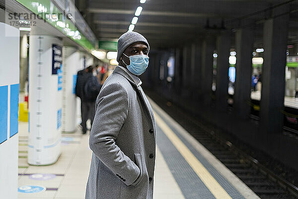 Geschäftsmann mit Hand in der Tasche und schützender Gesichtsmaske steht am Bahnhof