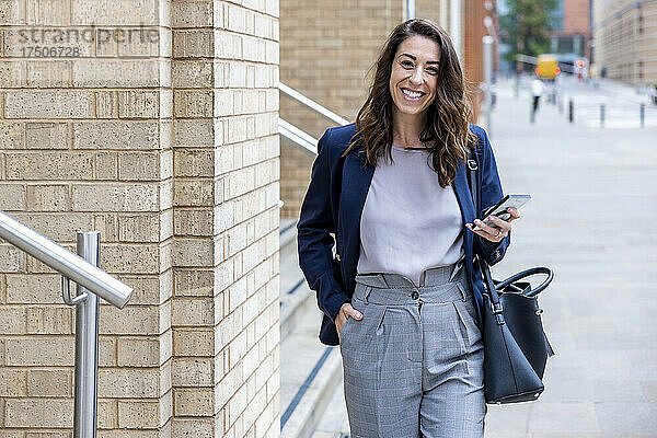 Lächelnde Geschäftsfrau mit der Hand in der Tasche und dem Mobiltelefon  die auf dem Fußweg läuft