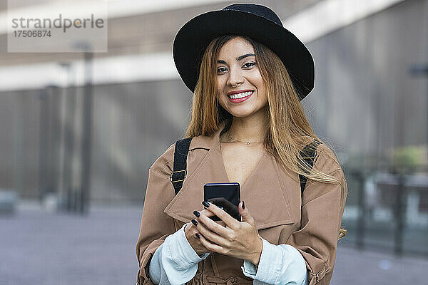 Frau mit Hut und Trenchcoat hält Smartphone in der Stadt