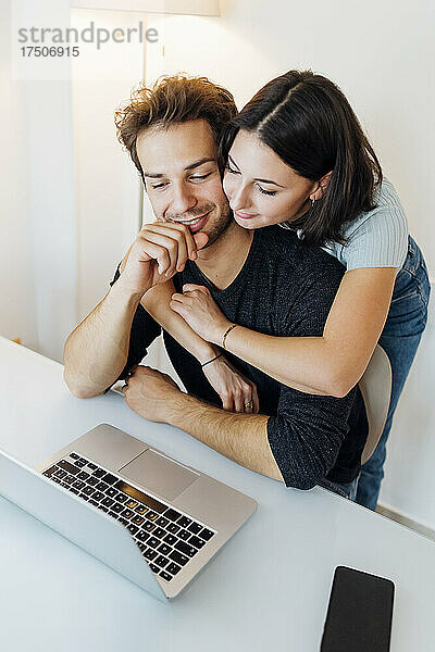 Junges Paar umarmt sich  während es zu Hause am Laptop arbeitet