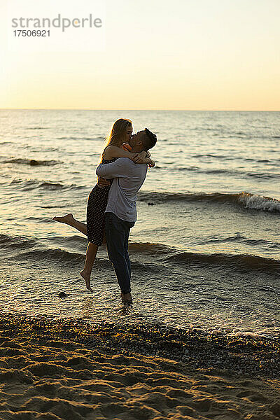 Romantischer Mann hebt und küsst Frau am Strand bei Sonnenuntergang