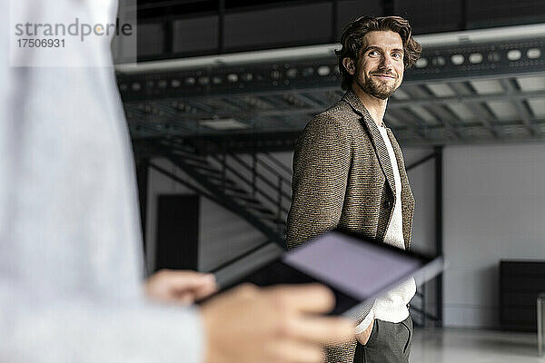 Lächelnder Geschäftsmann blickt Kollegen mit digitalem Tablet in Industriehalle an