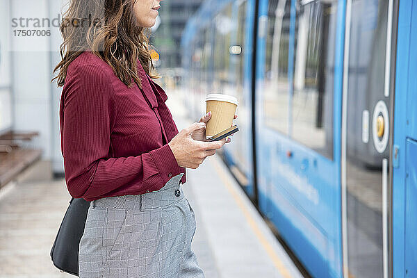 Geschäftsfrau mit Einwegbecher und Smartphone nähert sich am Bahnhof dem Zug