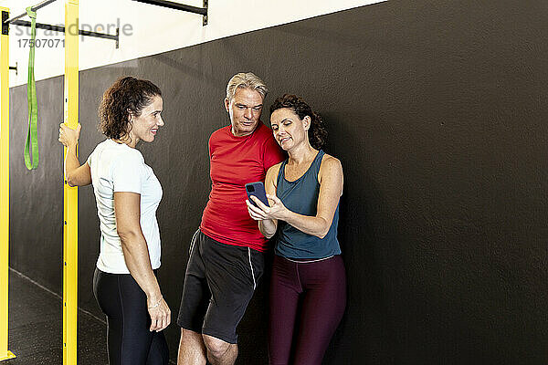 Lächelnder Athlet schaut Freunden zu  die im Fitnessstudio ihr Mobiltelefon teilen