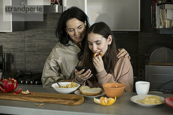 Mutter und Tochter schauen beim Frühstück in der Küche auf ihr Smartphone