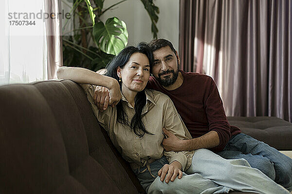 Lächelnder Mann umarmt Frau  die zu Hause auf dem Sofa im Wohnzimmer sitzt