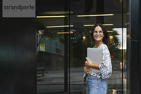 Lächelnde junge Geschäftsfrau hält Laptop vor dem Büro