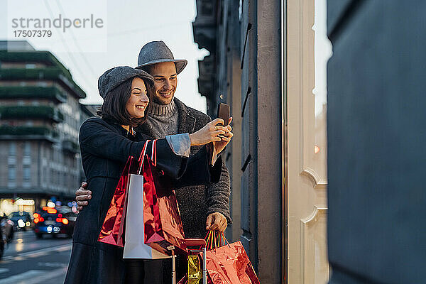 Lächelndes junges Paar mit Einkaufstüten  das in der Stadt ein Selfie mit dem Smartphone macht