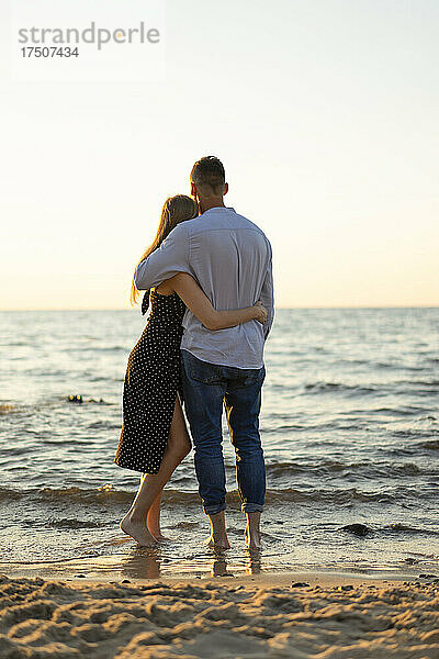 Romantisches junges Paar mit Armen und Blick auf den Horizont vom Strand aus