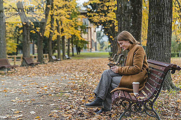 Lächelnde Frau benutzt Mobiltelefon auf Bank im Herbstpark