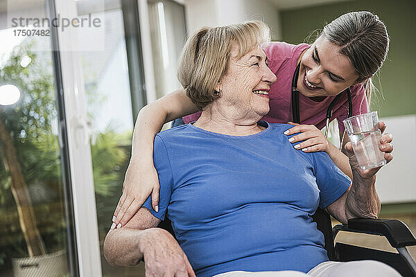 Lächelnde Pflegekraft steht zu Hause hinter behinderter Frau im Rollstuhl