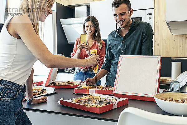 Lächelnde Geschäftsfrau fotografiert Kollegen  der in der Küche Essig auf Pizza gießt