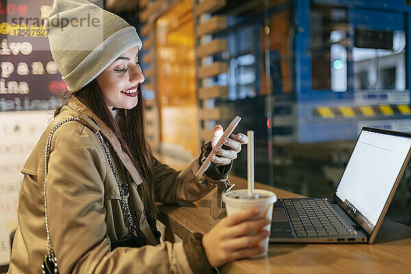 Frau benutzt Smartphone und hält Getränk am Laptop im Café