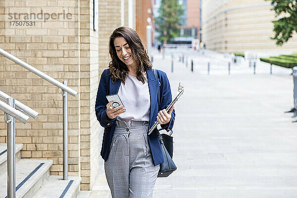 Lächelnde Geschäftsfrau mit Laptop und Smartphone geht auf Fußweg