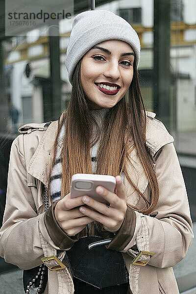 Lächelnde Frau mit Smartphone lehnt an Glaswand