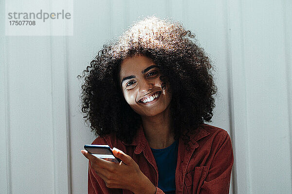 Lächelnde Frau mit Smartphone lehnt an weißer Wand