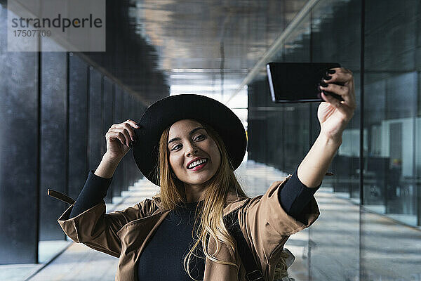 Lächelnde Frau mit Hut macht ein Selfie mit dem Handy auf dem Fußweg