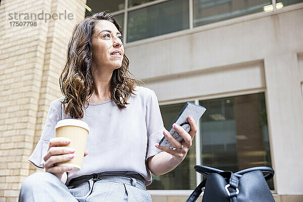 Nachdenkliche Geschäftsfrau mit Einwegbecher und Mobiltelefon