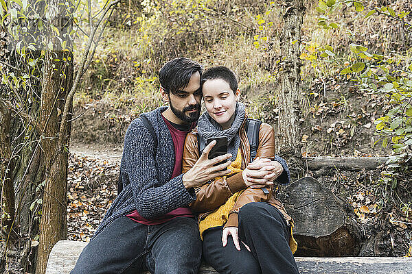 Bärtiger Mann teilt Smartphone mit Freundin auf Baumstamm am Flussufer