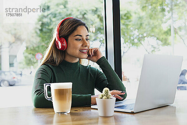 Lächelnde Geschäftsfrau mit Kopfhörern und Tasse arbeitet am Laptop im Café