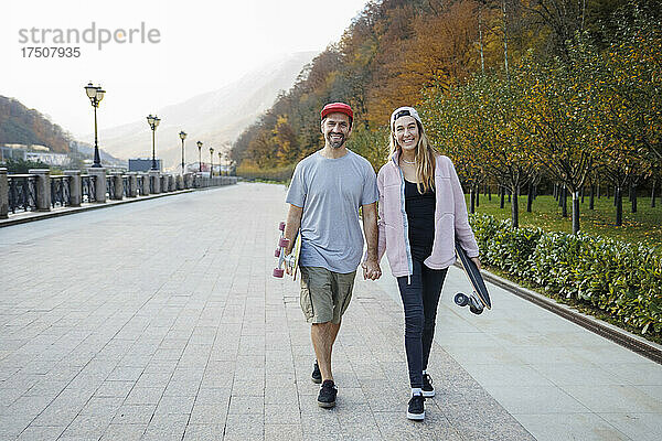 Lächelndes Paar mit Skateboards  die Händchen haltend gemeinsam auf dem Fußweg spazieren gehen