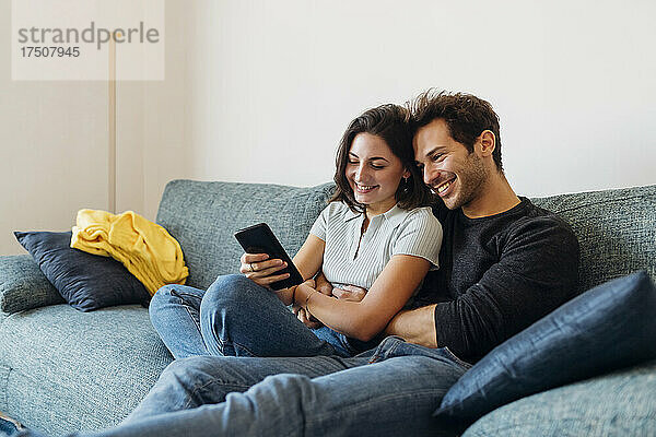 Lächelnde Freundin teilt Smartphone mit Freund auf Sofa zu Hause