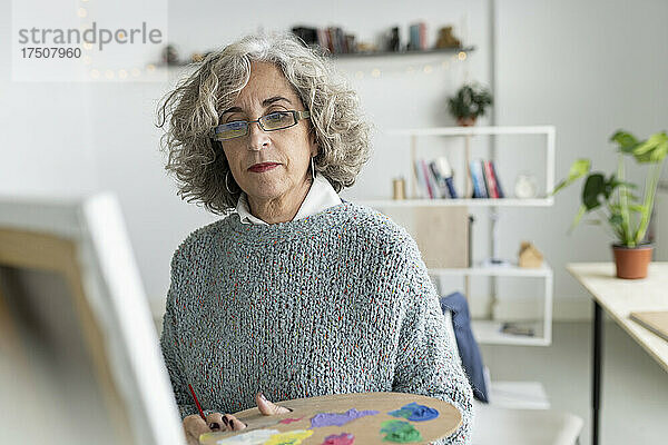 Ältere Frau mit Farbpalette und Blick auf Leinwand im Wohnzimmer