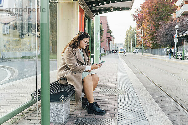 Üppige Frau benutzt Mobiltelefon  während sie an der Straßenbahnhaltestelle wartet
