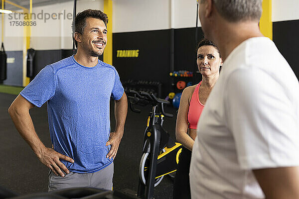 Lächelnder Sportler mit der Hand auf der Hüfte im Gespräch mit Freunden im Fitnessstudio